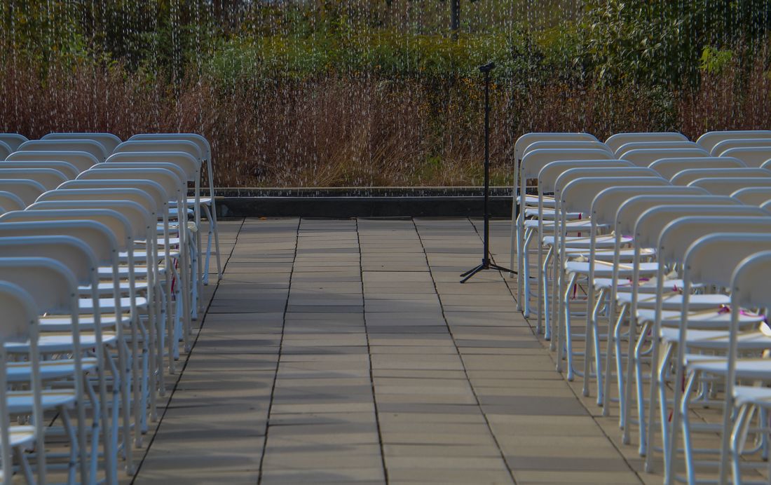 Rows of empty white folding chairs set up outdoors for a ceremony, with rain visible falling in the background.