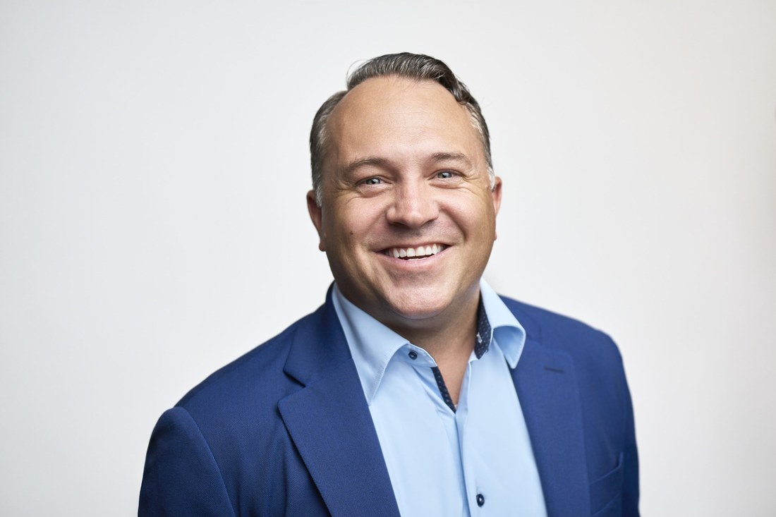 A smiling man in a navy blazer and open-collar light blue shirt, photographed against a plain studio backdrop for a corporate headshot