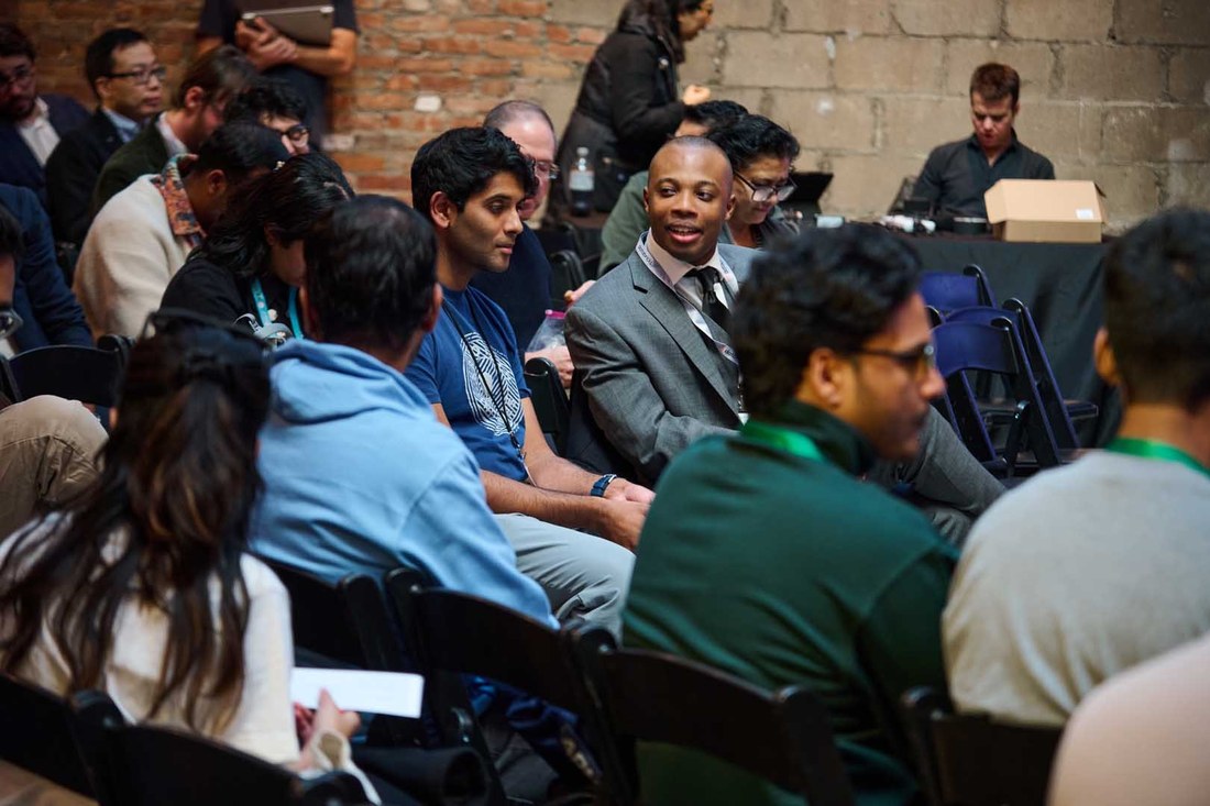 Conference attendees seated in folding chairs at a brick-walled event venue, talking with each other before the next session