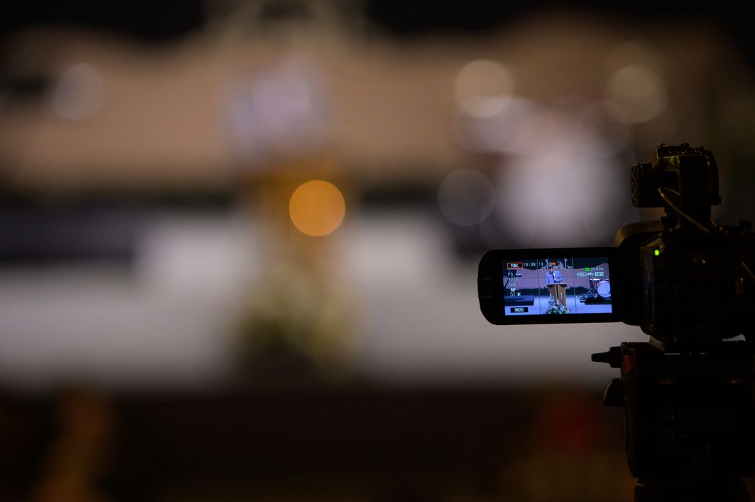 A video camera's flip-out screen shows a live event feed, with blurred string lights and a stage in the ambient background at night.