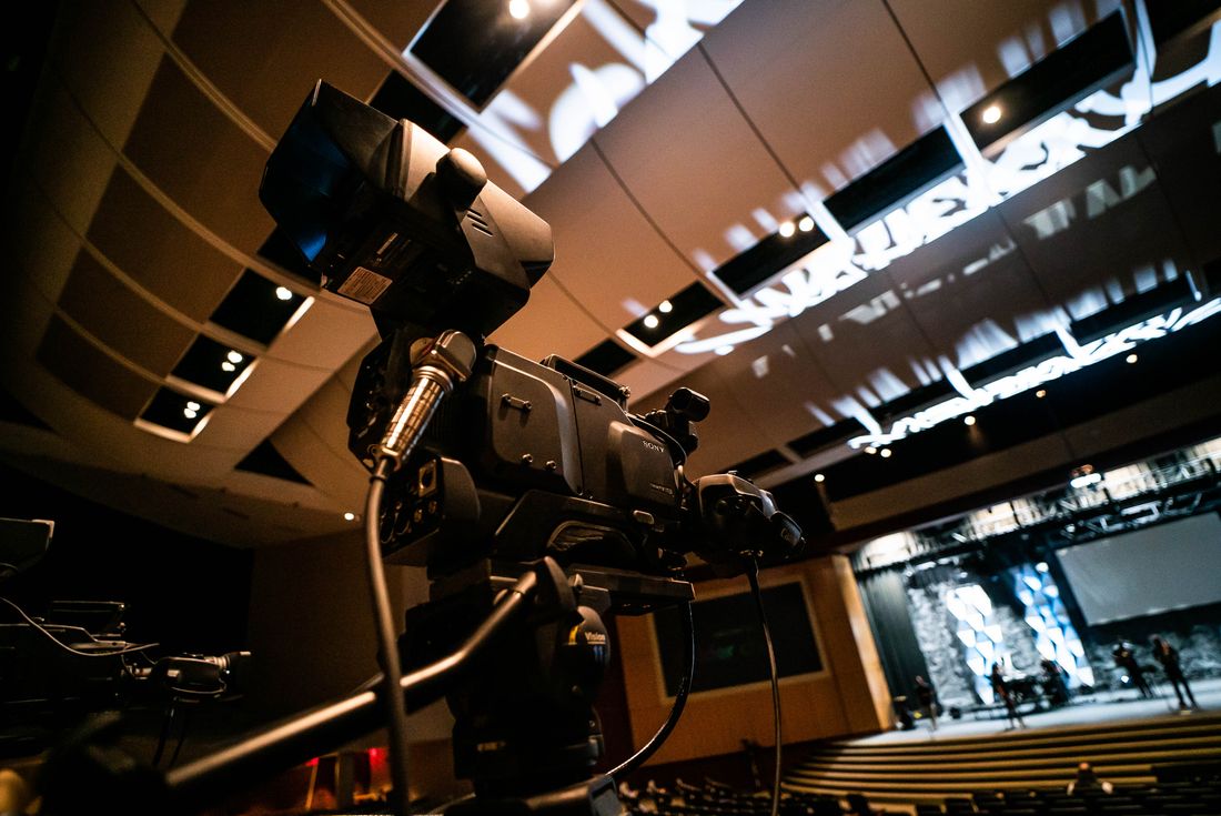 Professional video camera set up on a tripod in an auditorium, positioned for a wide static capture of the stage