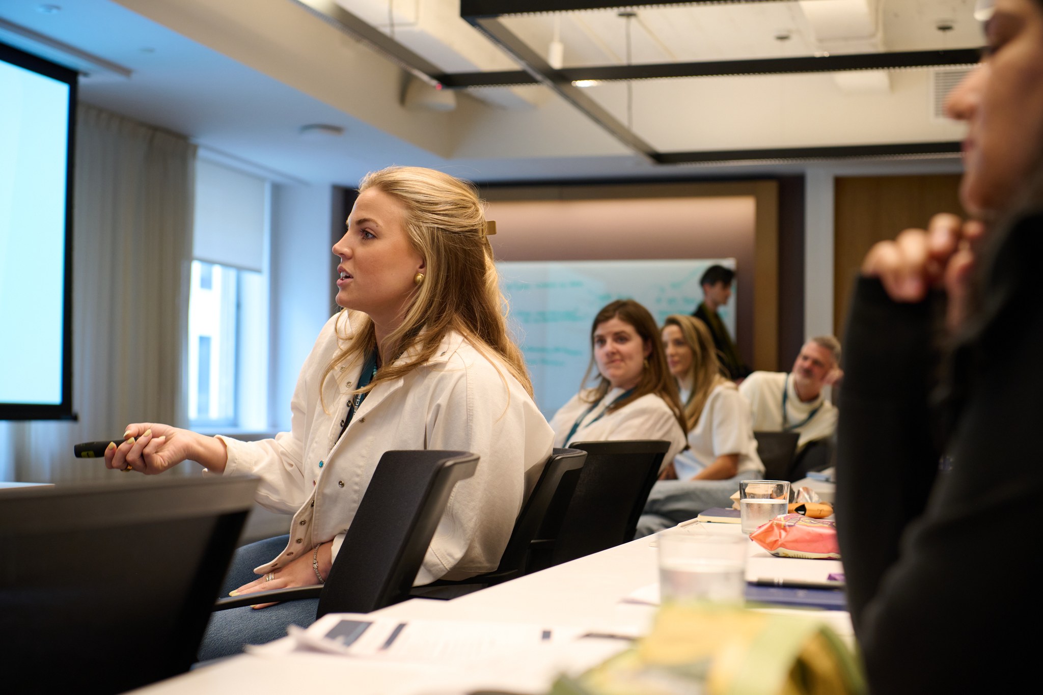 A woman with blonde hair speaks and gestures with a presentation clicker while seated at a conference table, with colleagues listening in the background.
