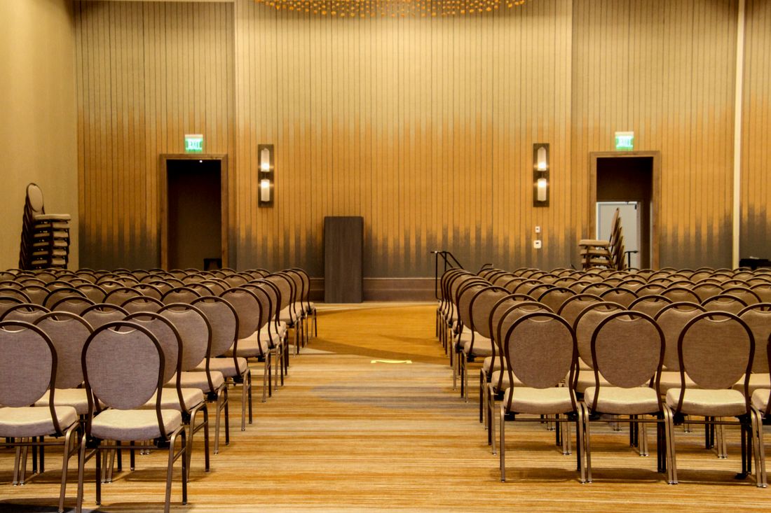 Rows of empty upholstered chairs facing a plain podium in a hotel conference room set up for a corporate event.
