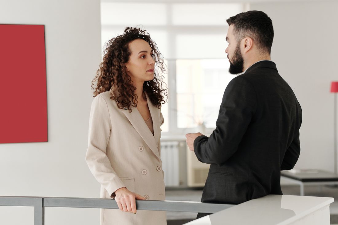 Two colleagues in business attire stand talking seriously in a plain, bright office.