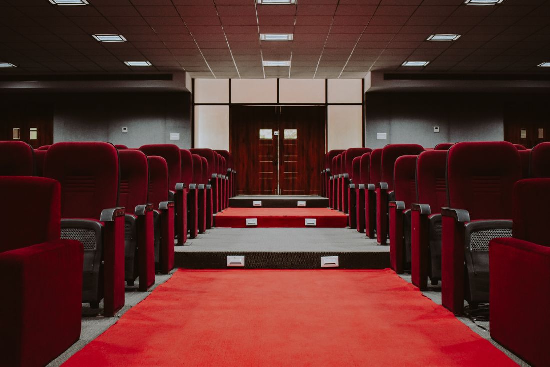 An empty theater-style auditorium with rows of red velvet seats and a wide red-carpeted center aisle leading to a small stage, no people present.