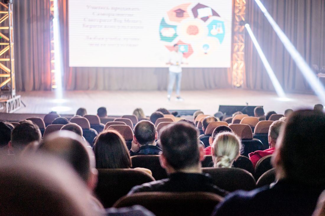 The backs of a seated conference audience in silhouette, facing a lit stage where a presenter stands beside a large screen under warm spotlight beams.