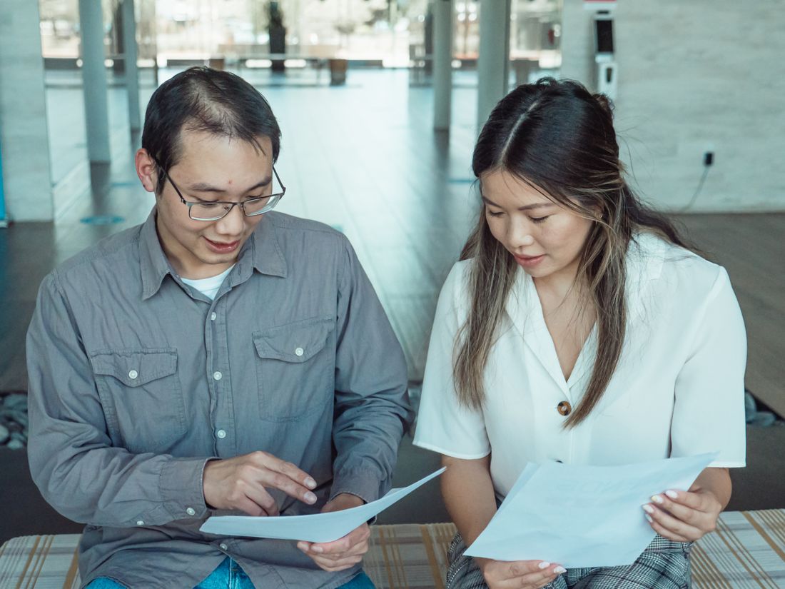 Two colleagues seated side by side reviewing printed pages, one pointing to a line on the page while the other reads her own sheet.