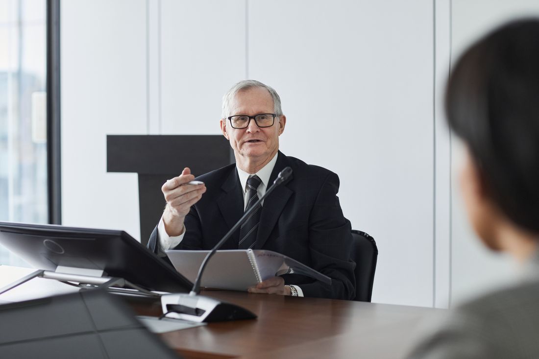 A senior man in a suit gestures mid-conversation at a conference room table, natural window light behind him and a monitor visible at the edge of frame.