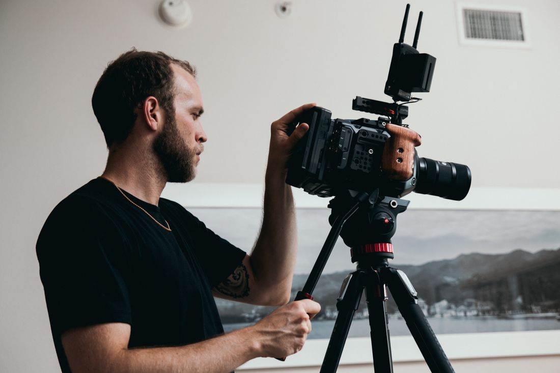 A crew member in profile adjusts a cinema camera mounted on a tripod in a plain, minimally lit room, no subject in frame.