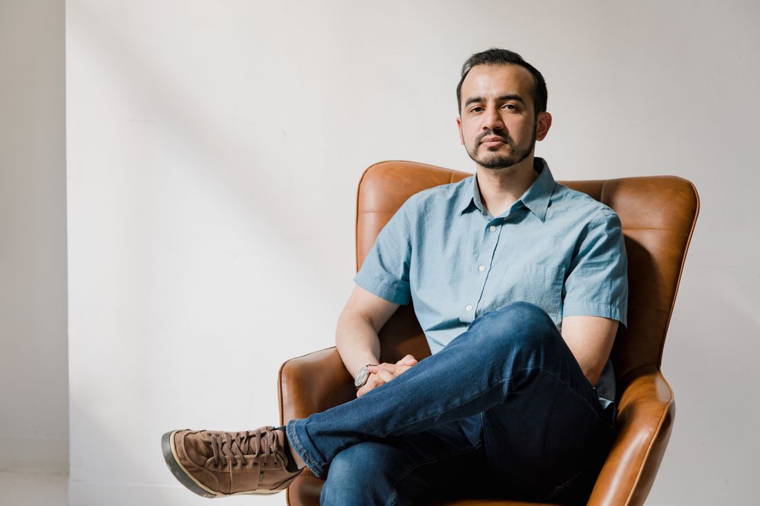 A man sits in a brown leather armchair against a plain white wall, wearing a light blue short-sleeve button-up shirt and jeans, looking toward the camera with a calm, relaxed expression.