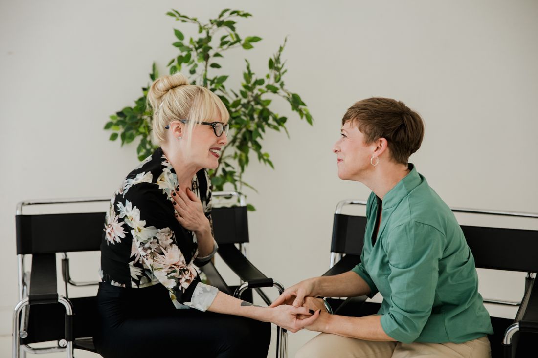 Two women sit facing each other in simple black chairs against a plain wall with a potted plant behind them, holding hands in warm conversation.