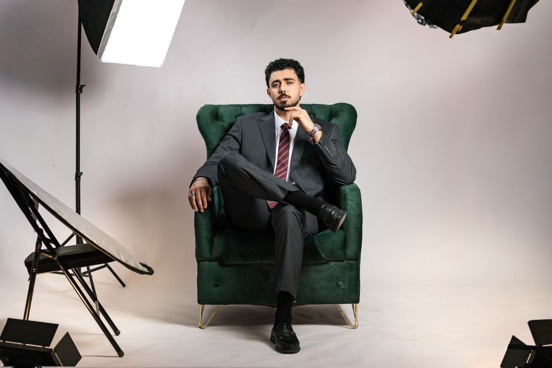 A man in a tailored suit sits for a portrait in a studio, softbox lighting visible in the frame.