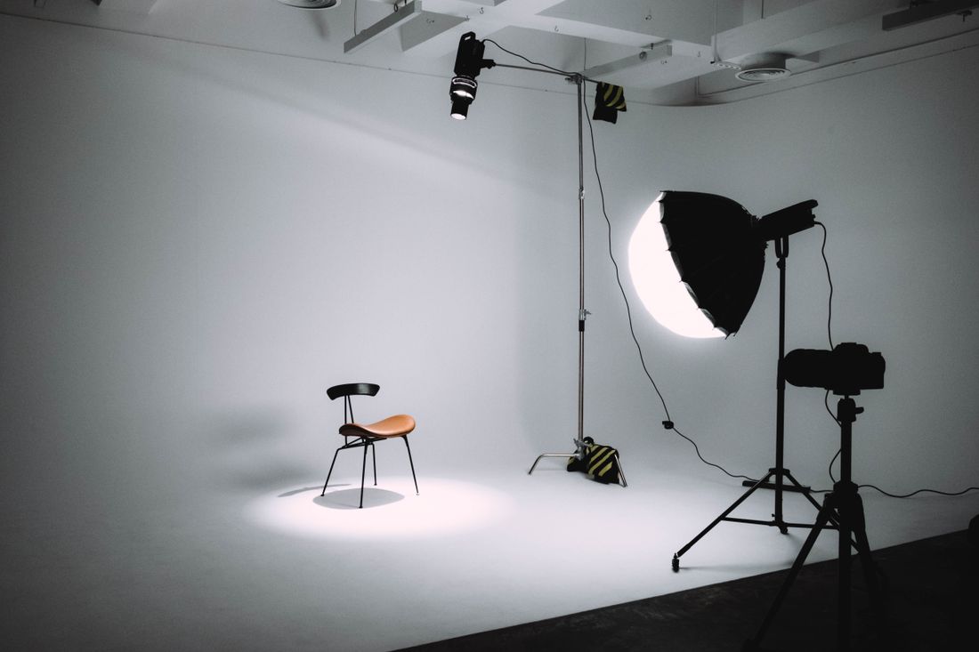 A single chair sits under a soft spotlight in an otherwise empty photography studio, camera ready on a tripod.