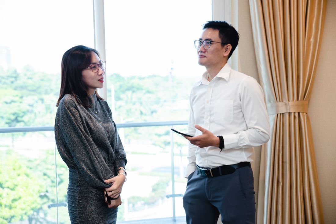 Two colleagues in business attire talk quietly near a large window with a curtain.