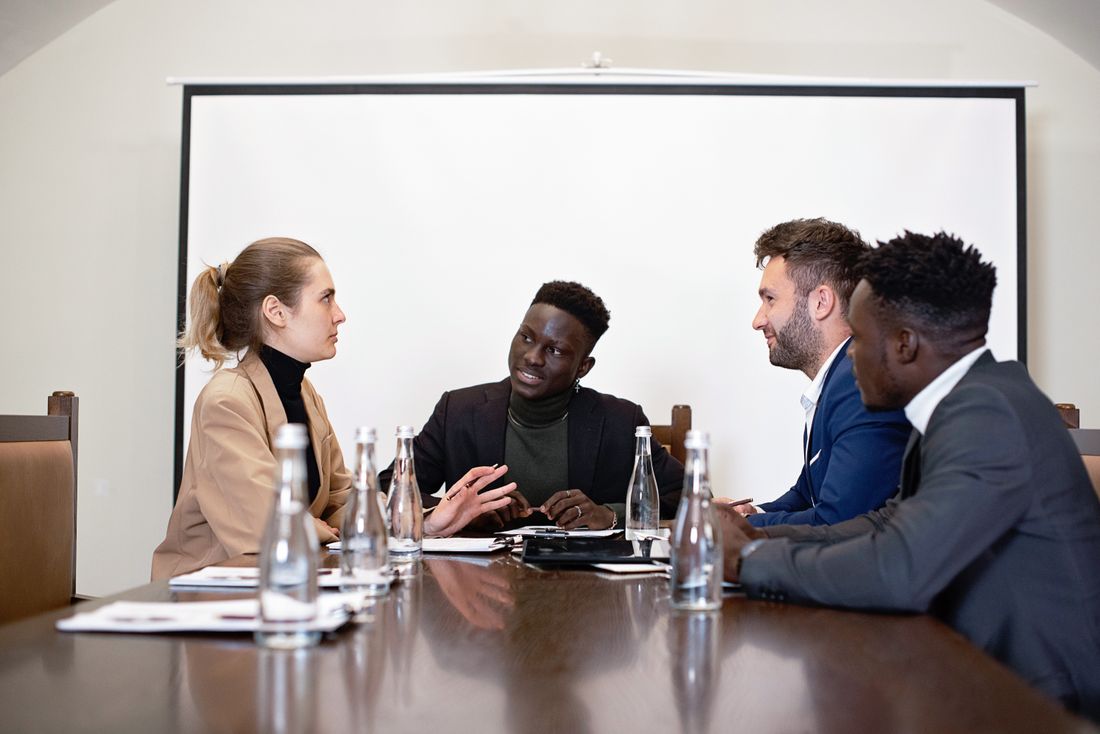 Four colleagues in business attire talk around a conference table with a blank screen behind them.