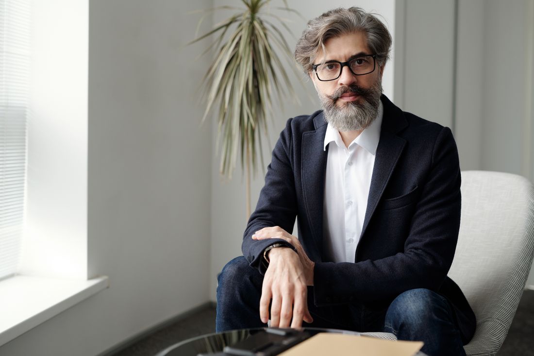 A gray-haired, bearded professional in a navy blazer sits near a window with soft daylight, a potted plant behind him, with a composed, steady expression.
