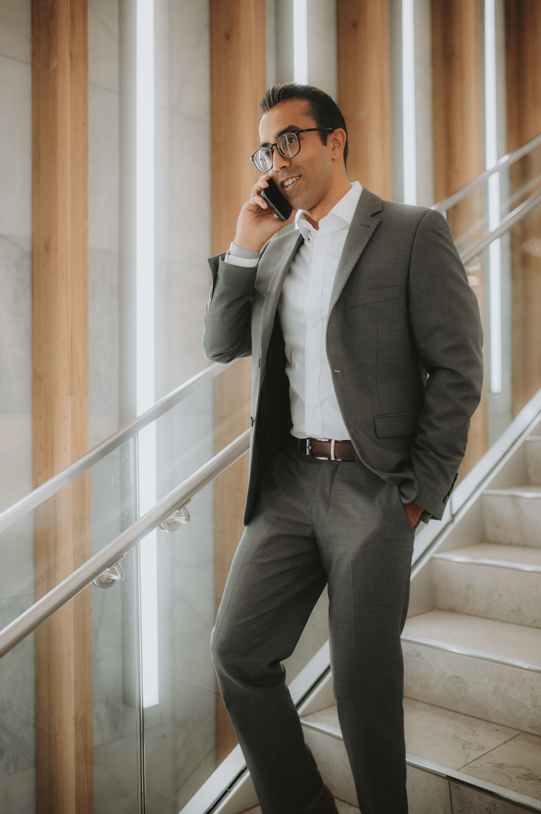A professional in a gray suit pauses mid-conversation on his phone while standing on an interior staircase with a glass railing and wood paneling.