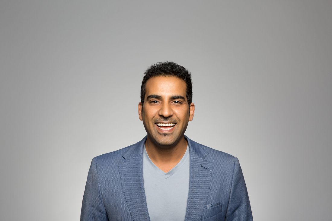 A man in a grey blazer over a v-neck shirt smiles warmly, photographed chest-up against a plain grey studio backdrop with even, soft lighting.