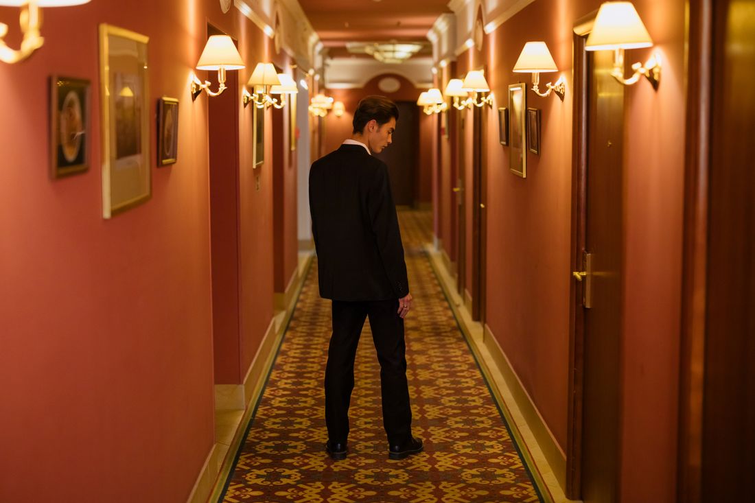 A man in a formal suit stands alone in an elegant hotel corridor lined with closed doors, suggesting a private transition between public moments.