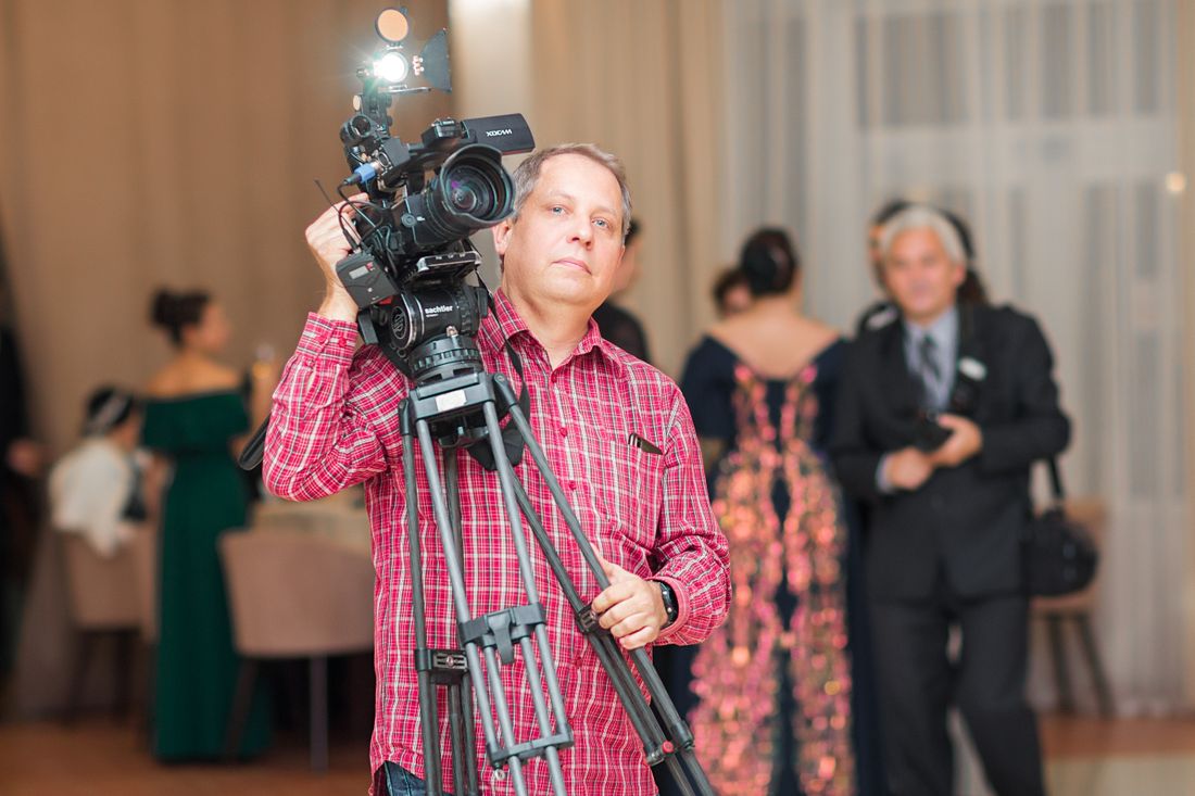 A photographer with a professional camera and tripod shoots discreetly from the side of a formal indoor event as guests in formalwear stand in the background.
