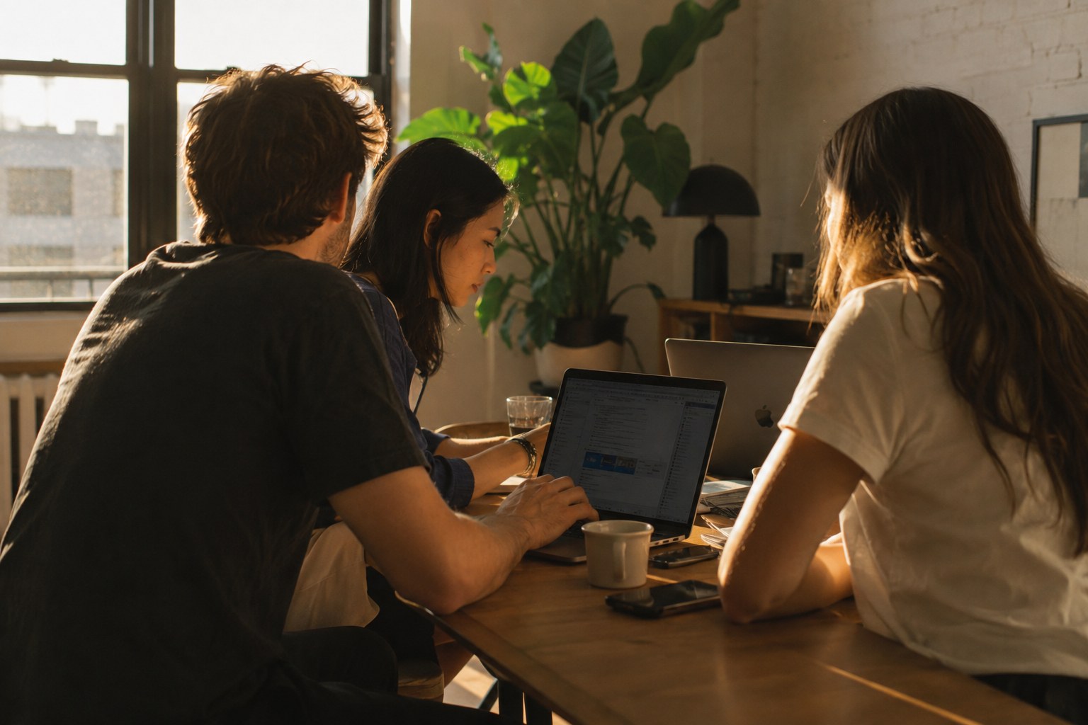 Three people collaborate around laptop at wooden table in warm golden light.