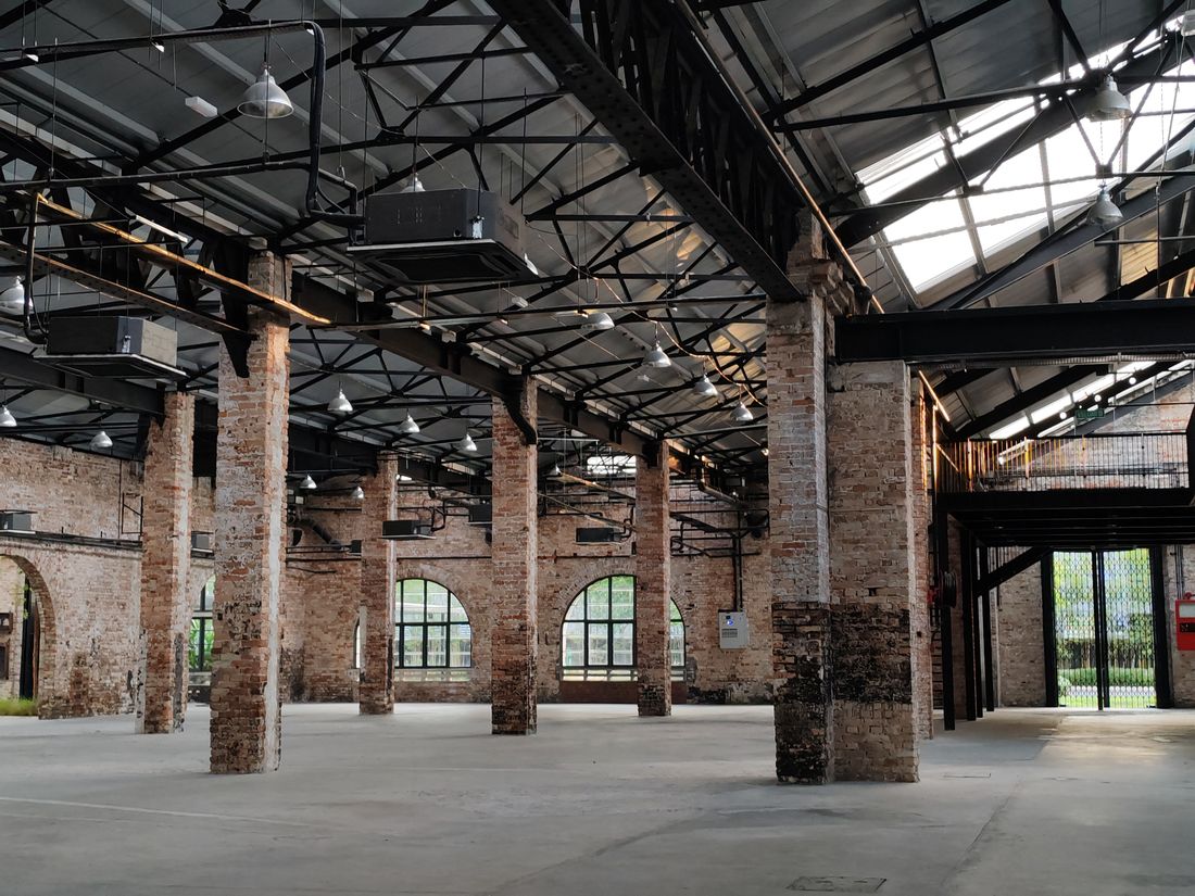 An empty, high-ceilinged industrial hall with exposed brick columns and arched windows, daylight streaming in through a glazed roof section.