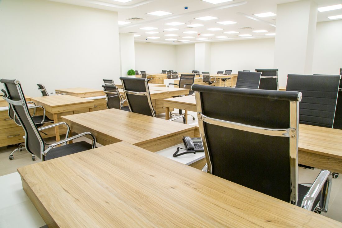 An empty open-plan office with light wooden desks, black office chairs and bright ceiling lighting.