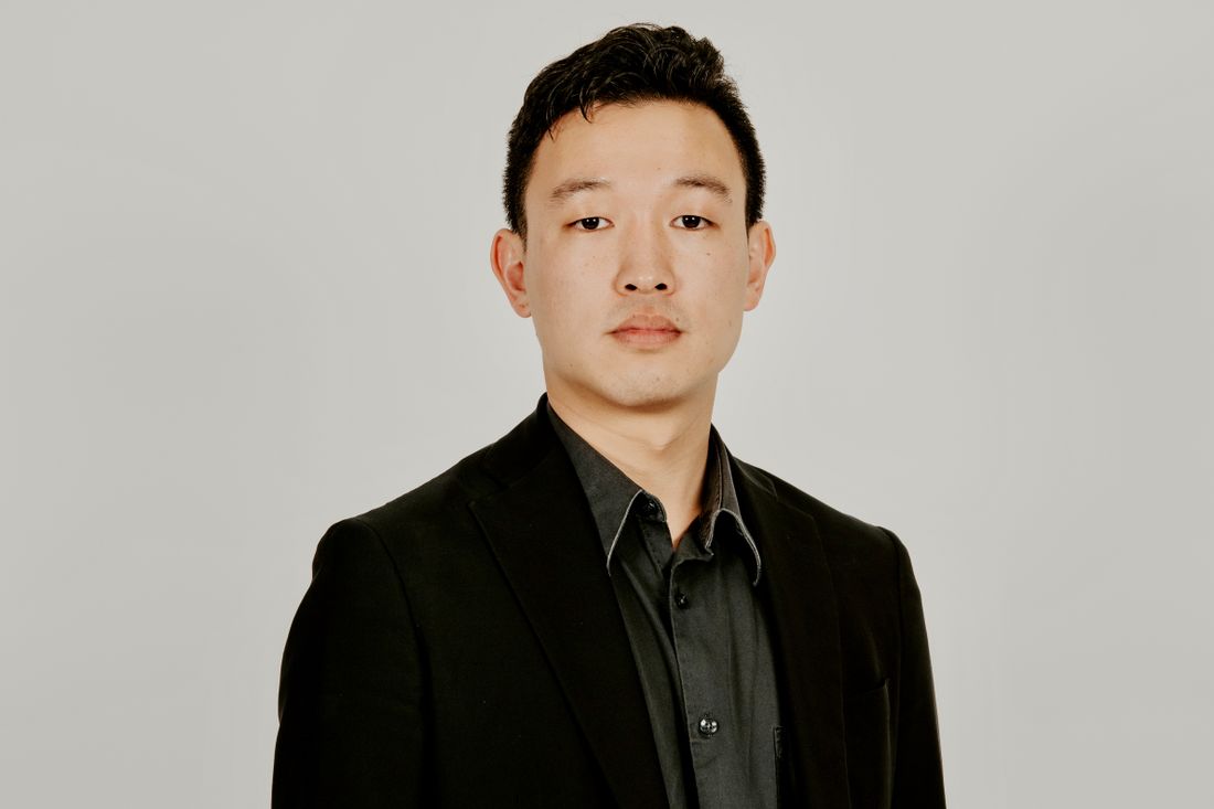 Chest-up studio portrait of a man in a dark suit and white shirt against a plain neutral gray background, calm and composed expression.