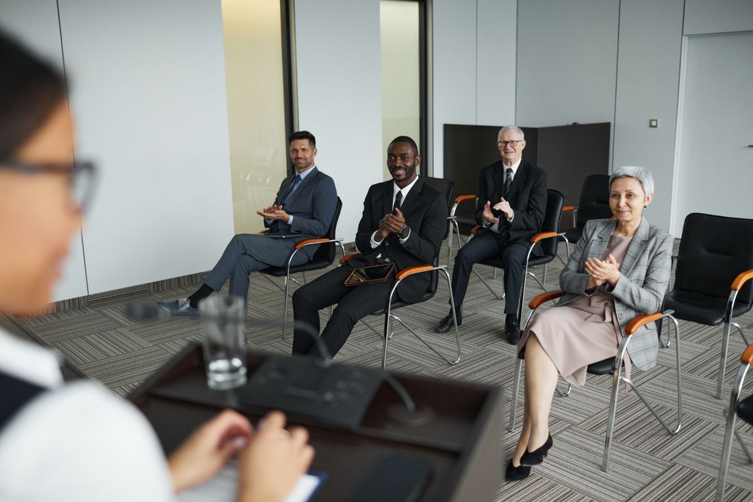 A small conference room of business-dressed attendees applauding while seated, with a presenter partially visible at a podium in the foreground, captured mid-recognition-moment.