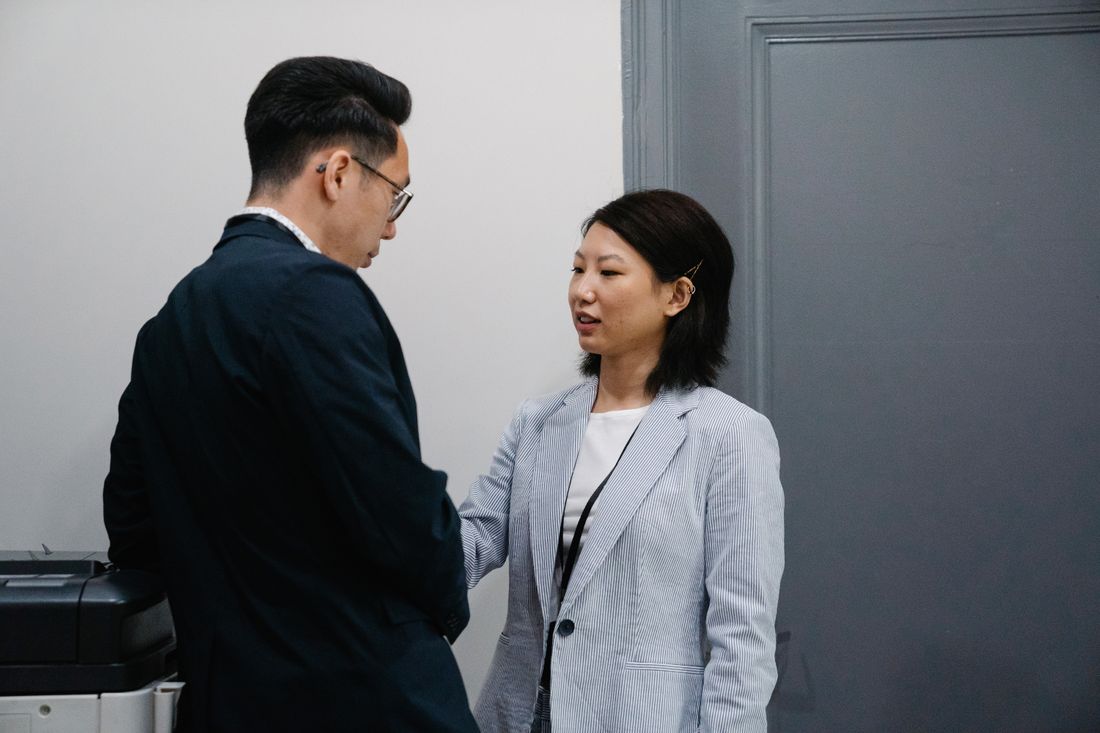 Two business-dressed professionals in an office hallway mid-conversation, greeting each other, candid unposed framing.