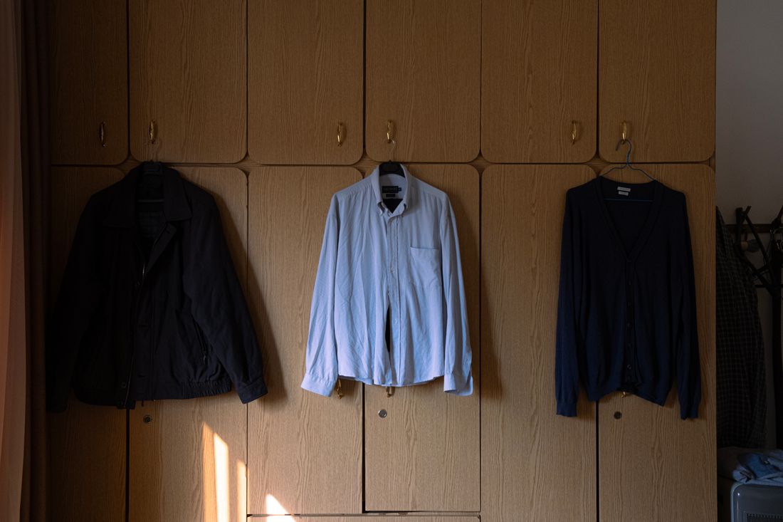 A jacket, a button-down shirt, and a cardigan hang from hooks on a plain wood cabinet wall, lit by low morning sunlight.