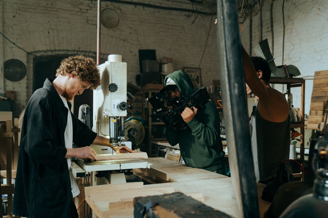 A craftsman works at a workshop machine while a camera operator films the process up close.