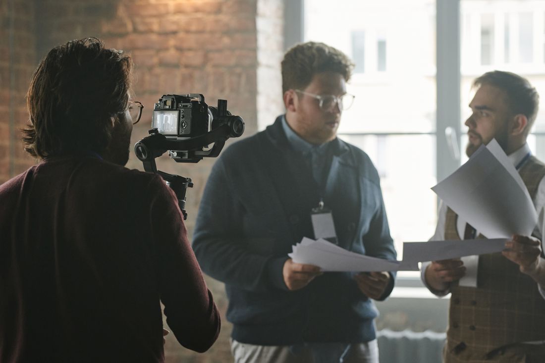 A camera operator films two colleagues reviewing papers together in a sunlit loft office.