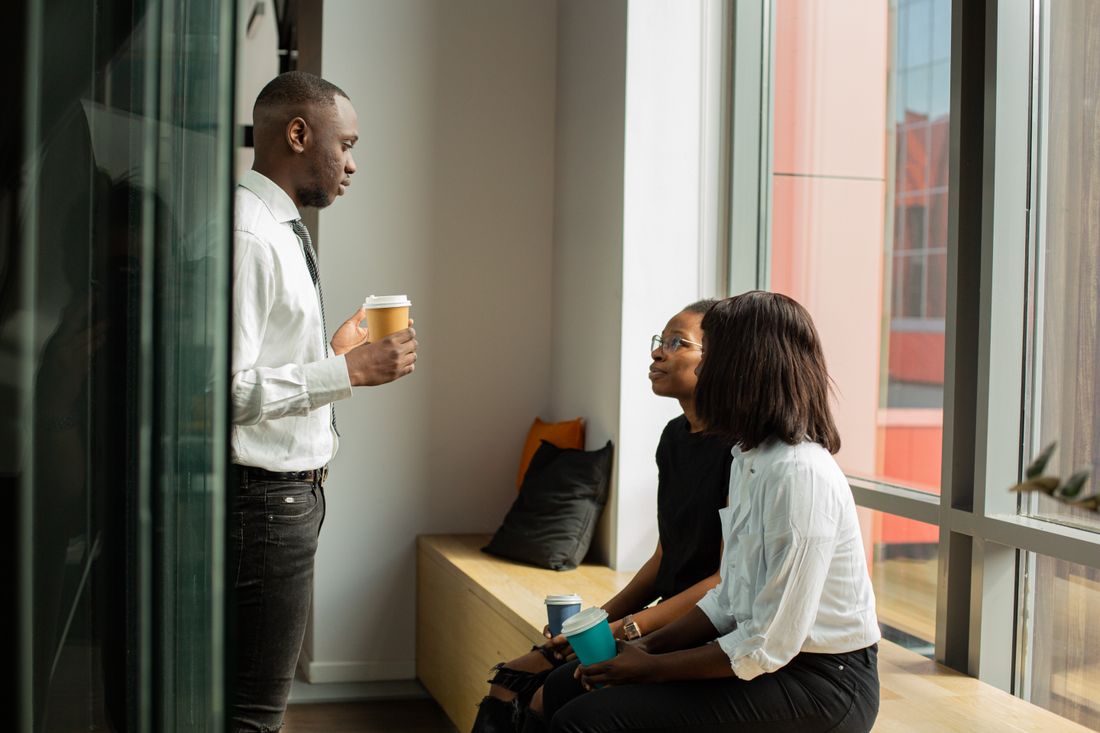 Three colleagues talking near a bright office window, one holding a coffee cup, in a casual mid-conversation moment.