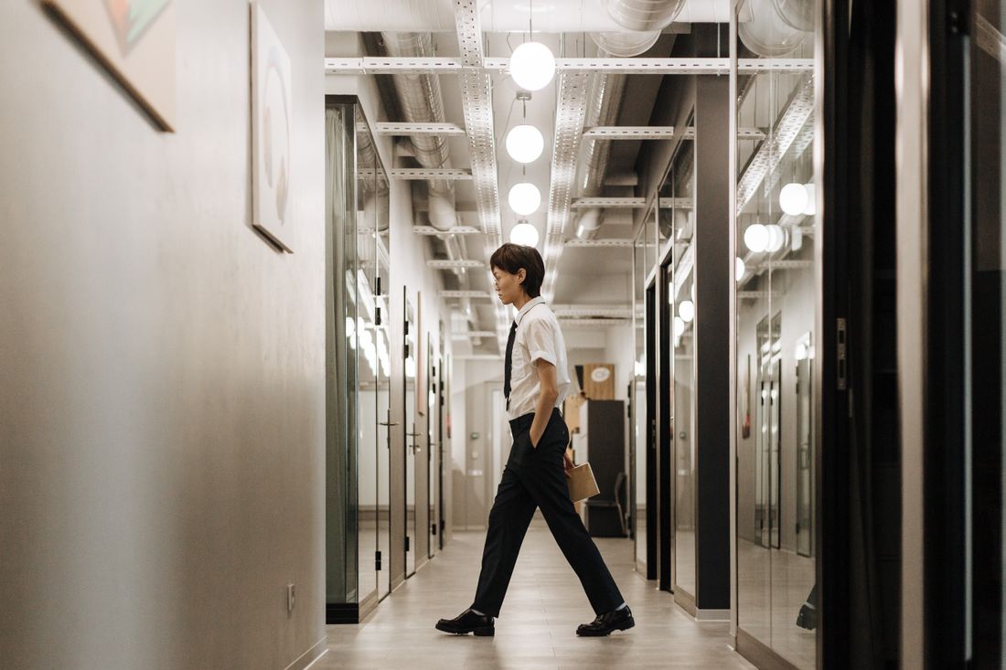 A person in business attire walking down a bright office corridor lined with glass-walled meeting rooms.