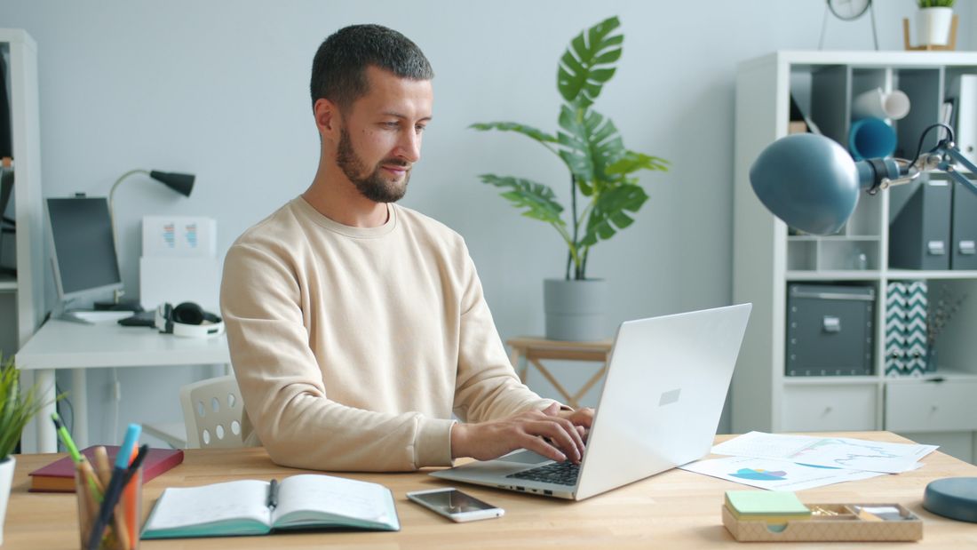 A man works alone at a laptop in a plain, plant-filled home office with open shelving and natural light.