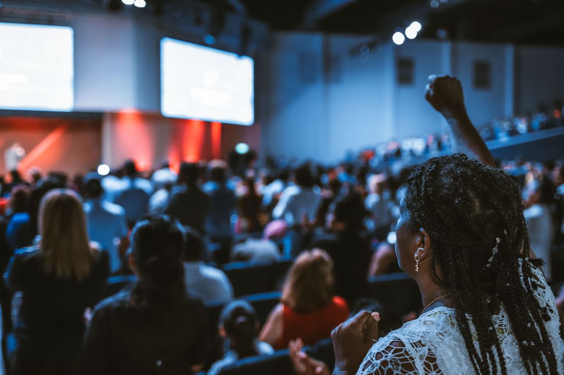 Wide view of a conference audience seen from behind, seated in a full room facing a blurred stage with warm lighting in the distance.