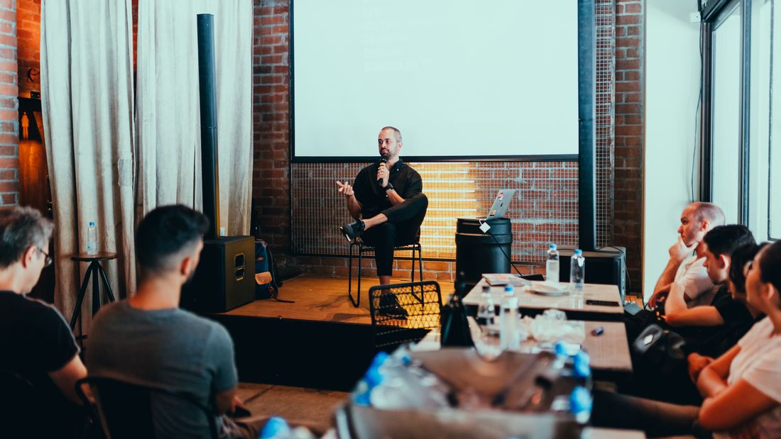 A presenter seated on a small stage holding a microphone and addressing a seated audience in an industrial-style conference room, with a blank projection screen behind him.