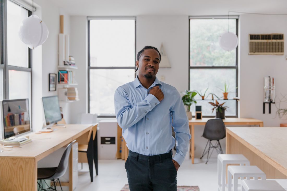 A man in a light blue button-down shirt stands in a bright, plain studio-style room with white walls and large windows, adjusting his collar.