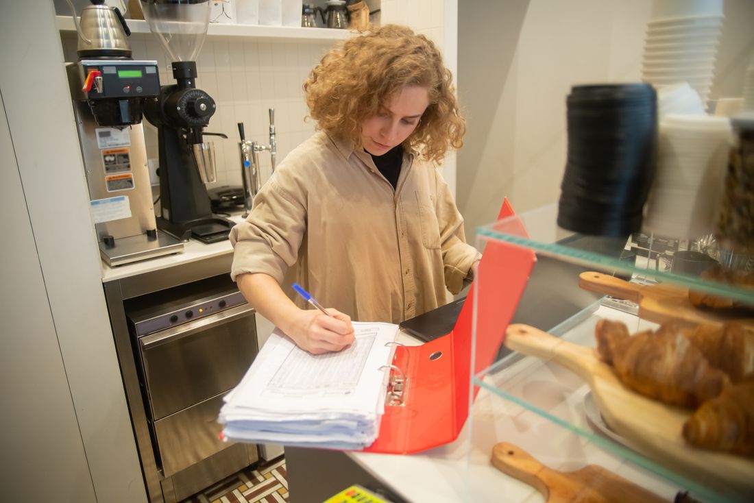 A woman in a small cafe writes on paperwork in a binder near an espresso grinder and a pastry display case, with counter equipment visible around her.