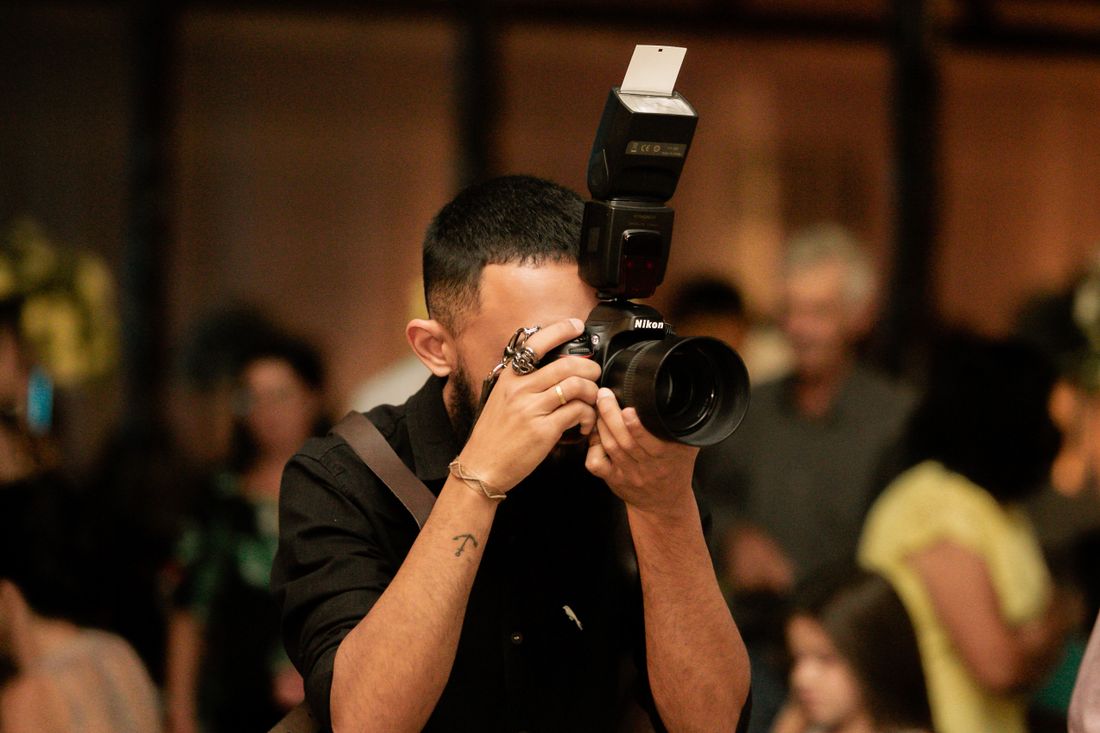 A photographer holds a camera with an on-camera flash unit up to shoot toward a crowded, warmly lit indoor event.
