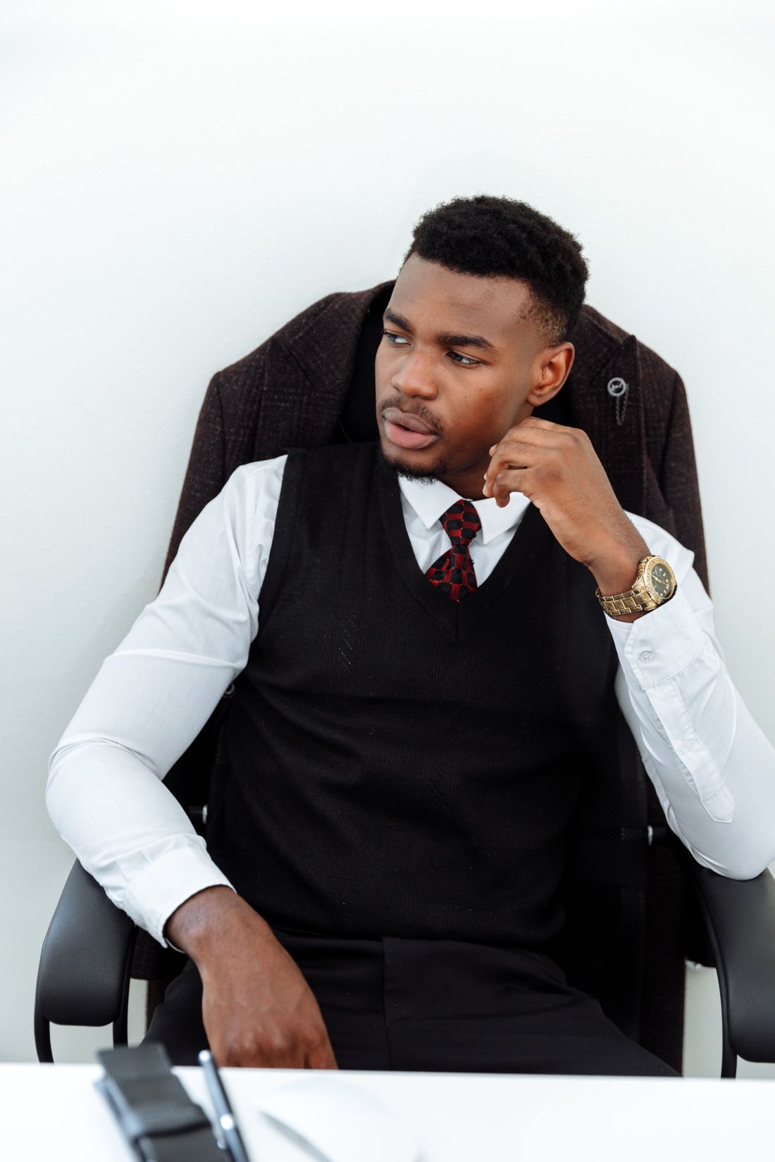 A composed professional in a vest and tie sits back in an office chair, hand resting near his chin, against a plain white wall.