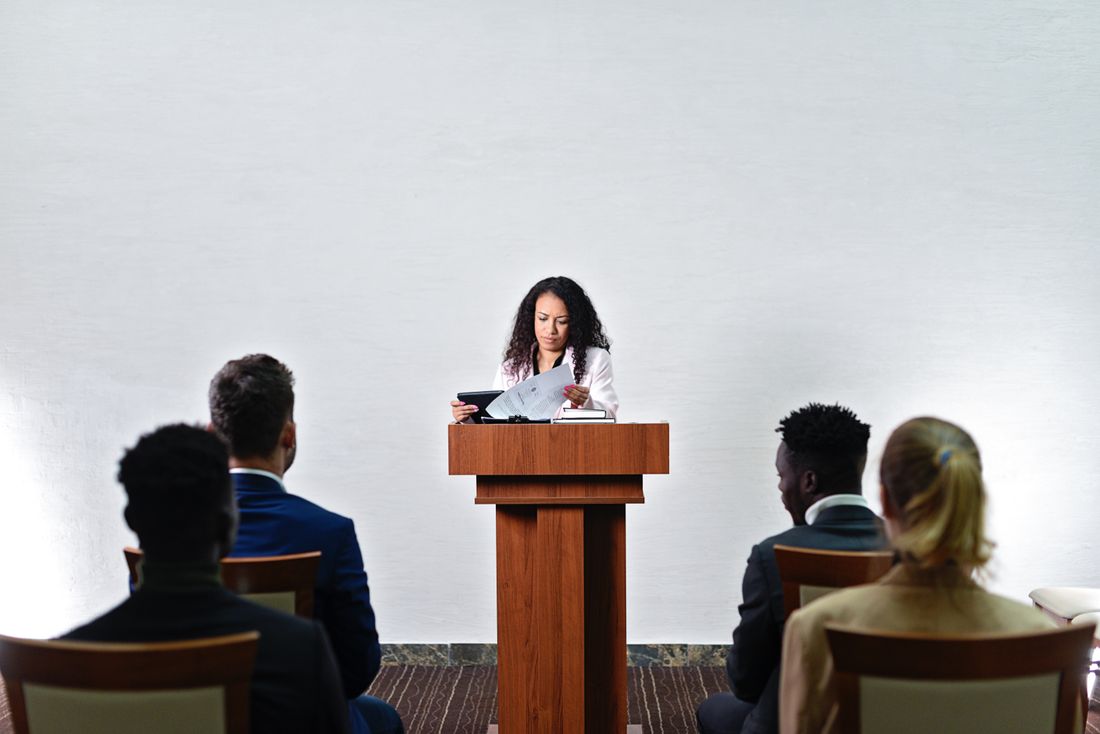 A speaker reading from papers at a plain wooden lectern against a bare white wall, seen from behind a small seated audience.