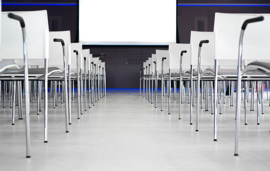 Rows of empty stackable chairs facing a blank projection screen in a plain, dimly lit conference room, seen from floor level at the back.