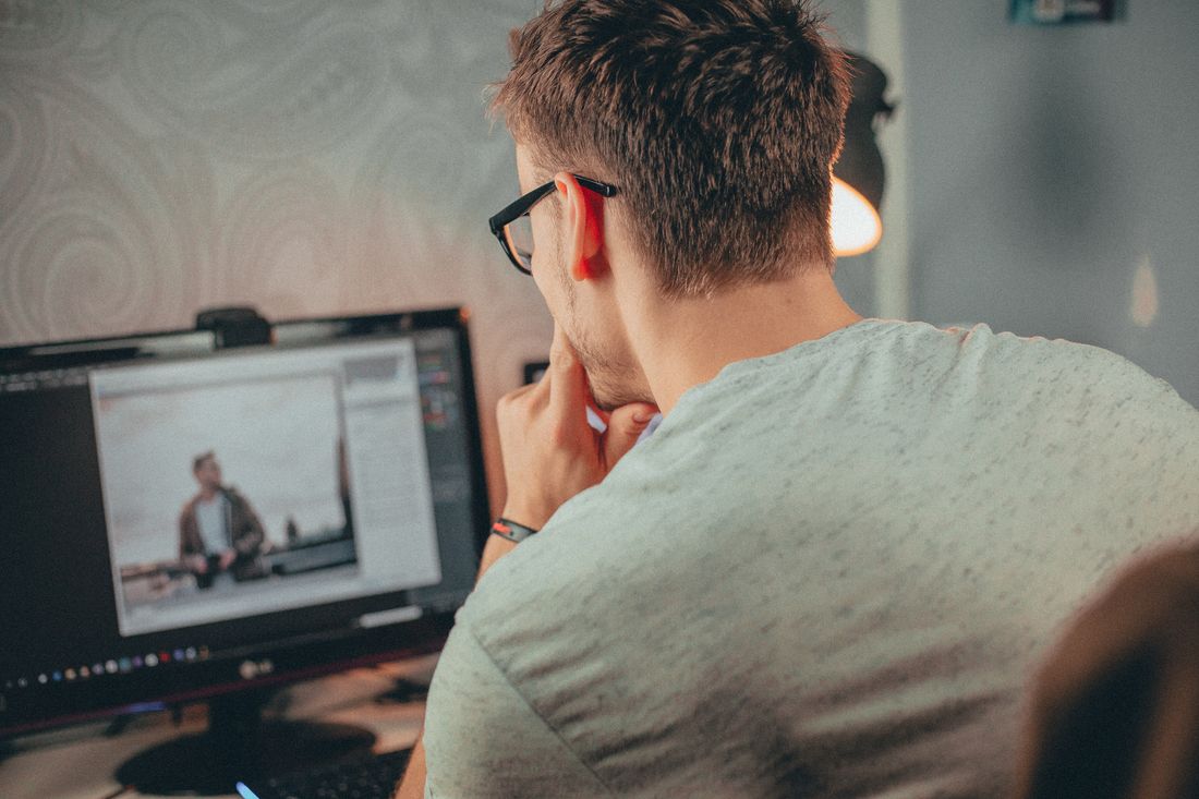 Seen from behind, a person wearing glasses studies a portrait photograph open in editing software on a desktop monitor in a dimly lit room.
