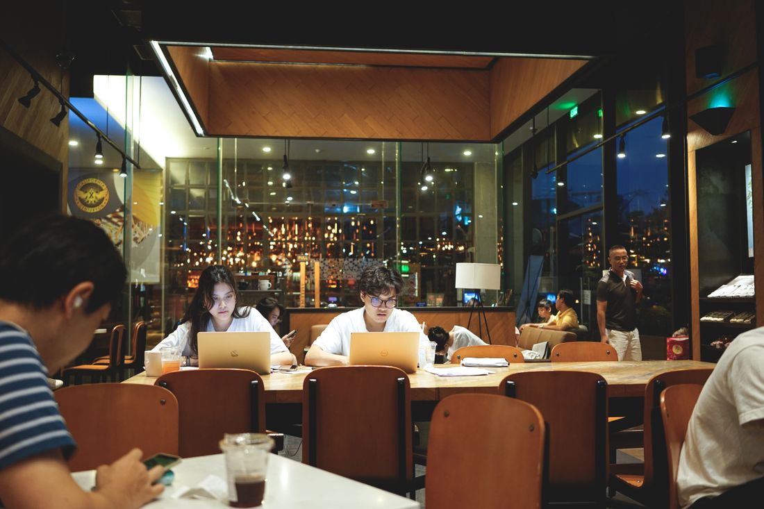 Two people at a long table late at night, heads down over open laptops under warm ambient lighting with a city view behind them.