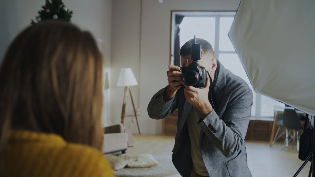 A photographer with a studio softbox photographing a portrait subject in a casual indoor event setting.