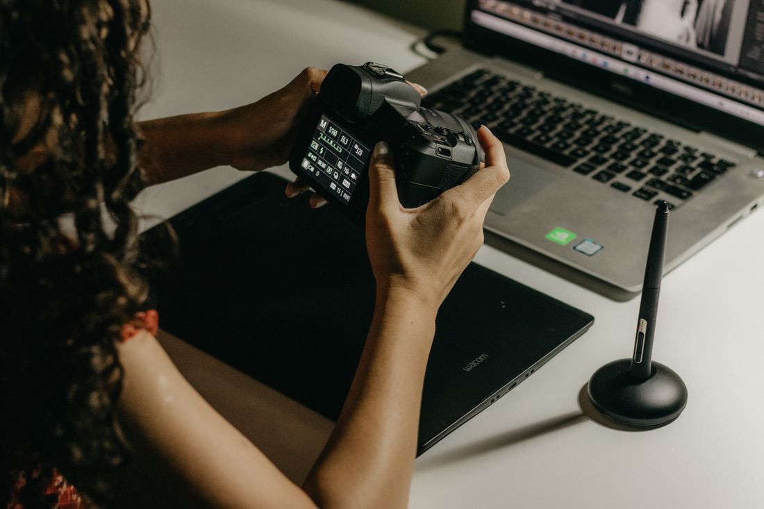 Close-up of hands holding a camera to review a shot next to a laptop and graphics tablet on a desk.