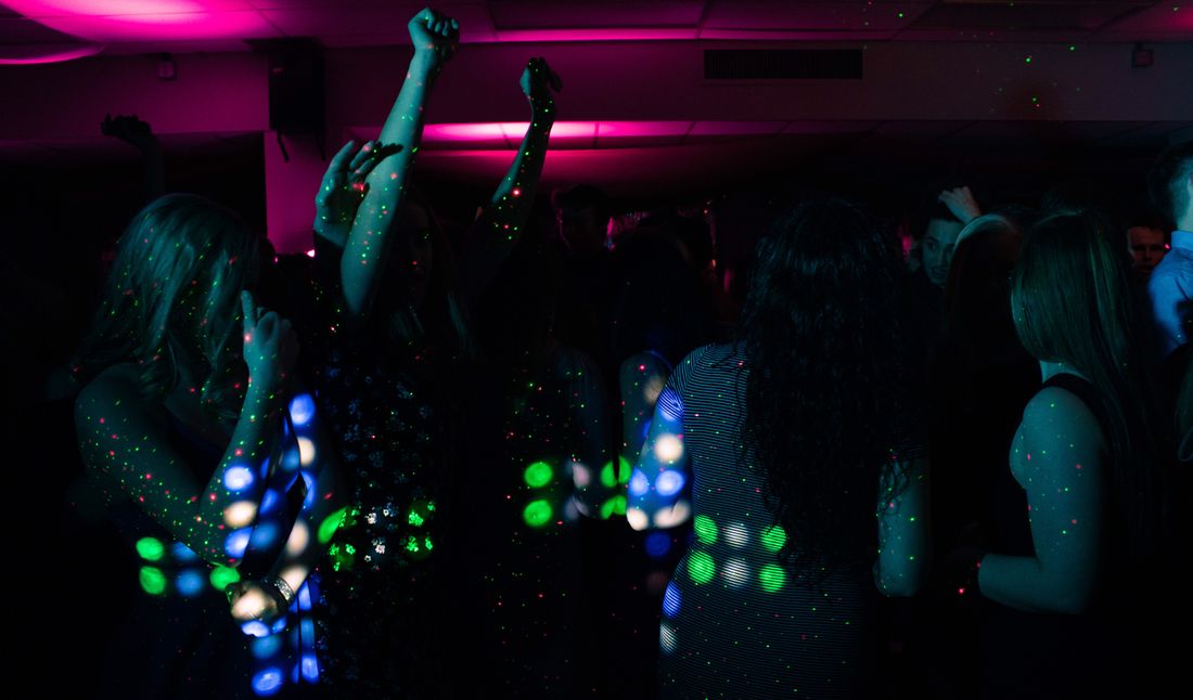 A group of people dancing with arms raised under pink and green colored party lighting, faces turned away or in shadow.