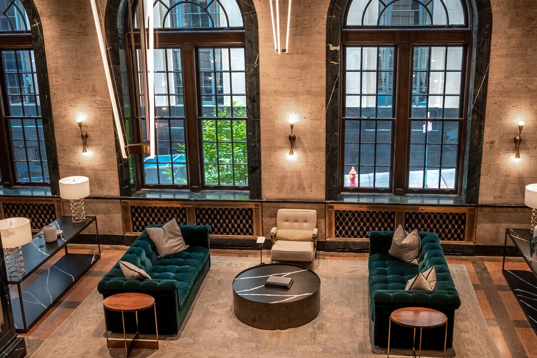 A hotel lobby seating group with dark green tufted sofas beneath tall arched windows, warm stone walls and brass sconces, no people in frame.